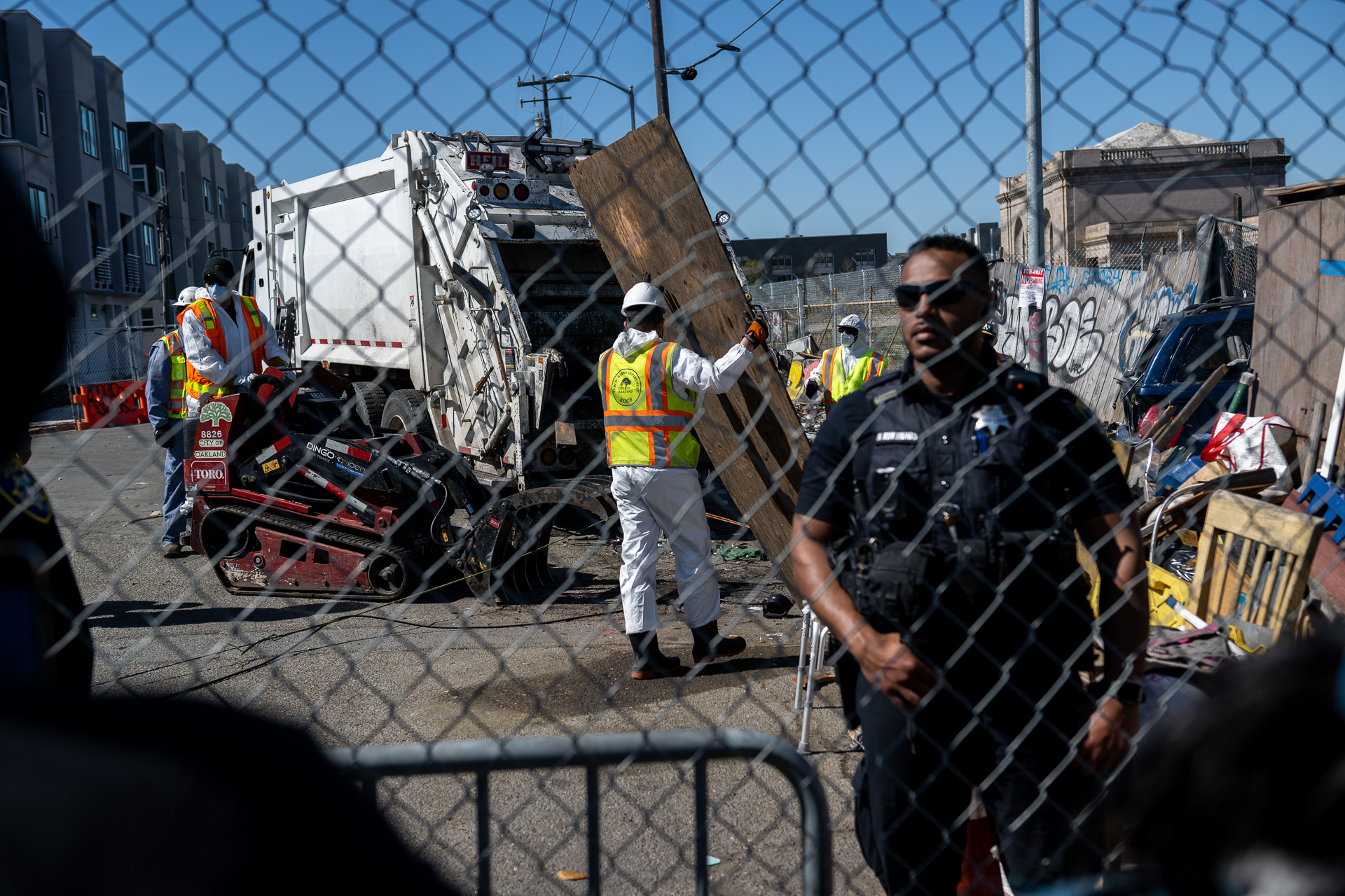 A Black police officer is shown standing behind a chain link fence as city workers behind him haul wood and other materials into large garbage trucks.