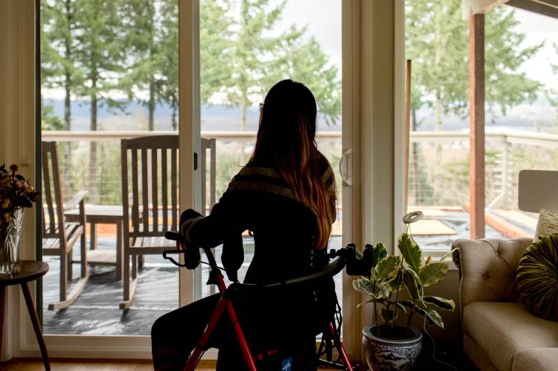 A woman, in shadow, with her back to the camera, sits on a rolling walker and stares out the window.