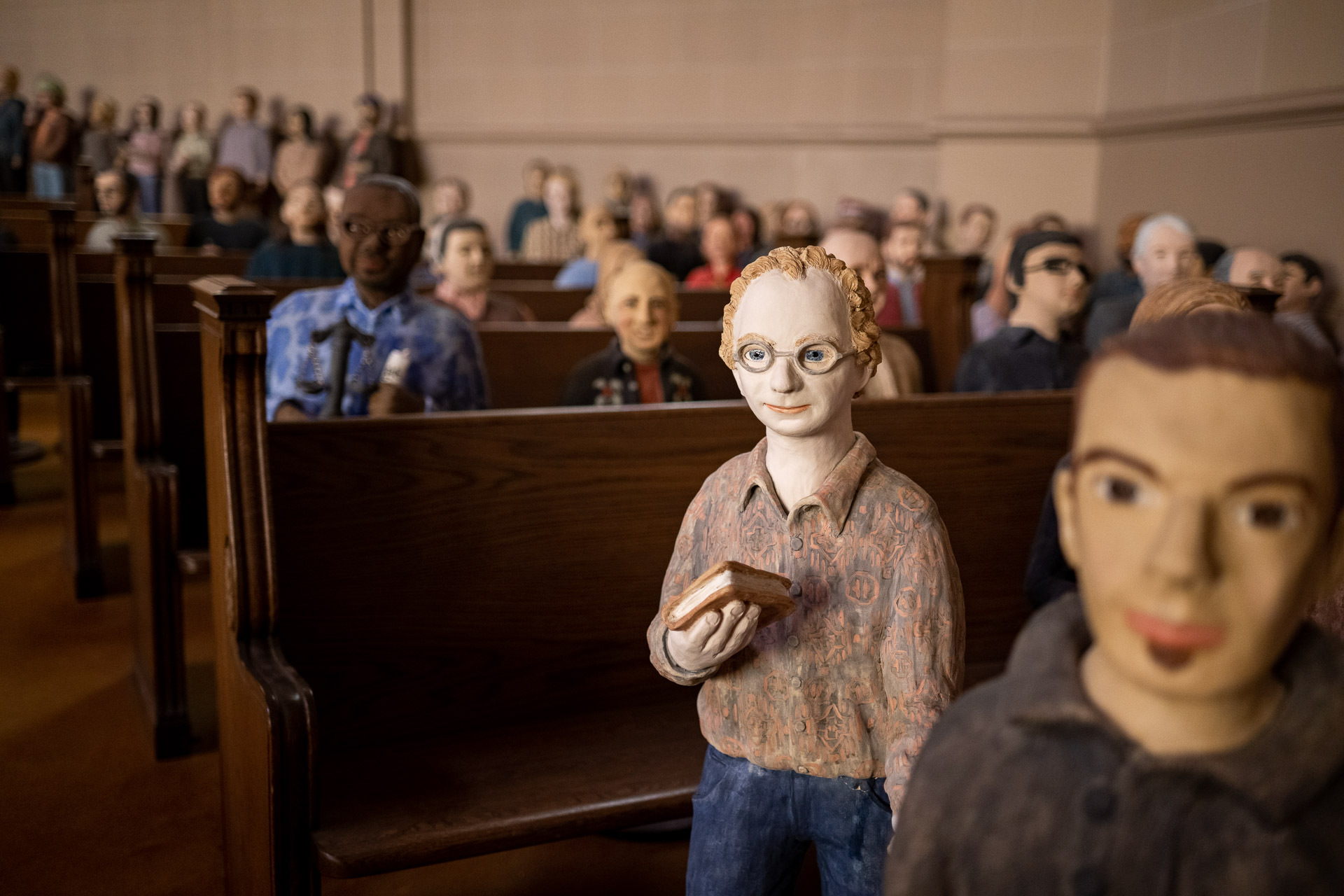 A photo of what appear to be dozens of clay figurines which are delicately painted and apparently standing near church pews
