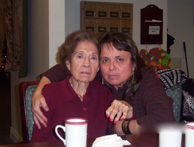 A photo of two women with the woman on the right with her arm over the other woman's shoulder while she is seated at a table with a cup in front of them.