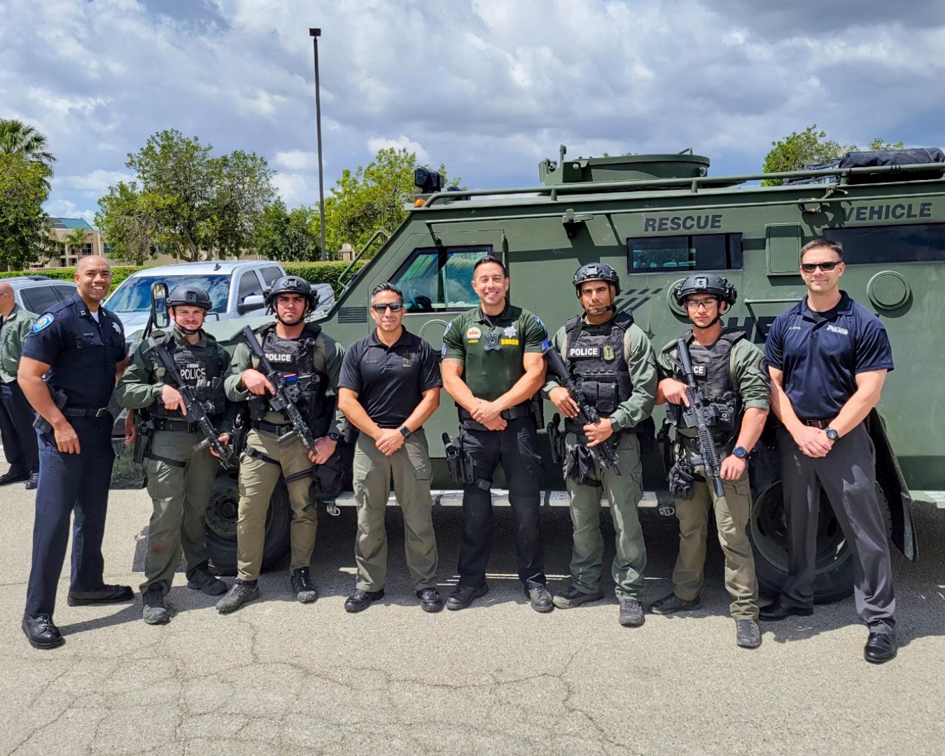 A police SWAT team poses in front of an Army green armored vehicle holding weapons.