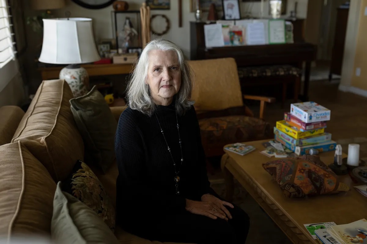 An older white woman with shoulder-length, gray hair and black outfit sits on a tan couch inside her home. A wooden coffee table is in front of the couch and holds stacks of board games, candles and random paperwork. A dark, wooden piano with sheet music is pictured in the background, along with photos on the walls.