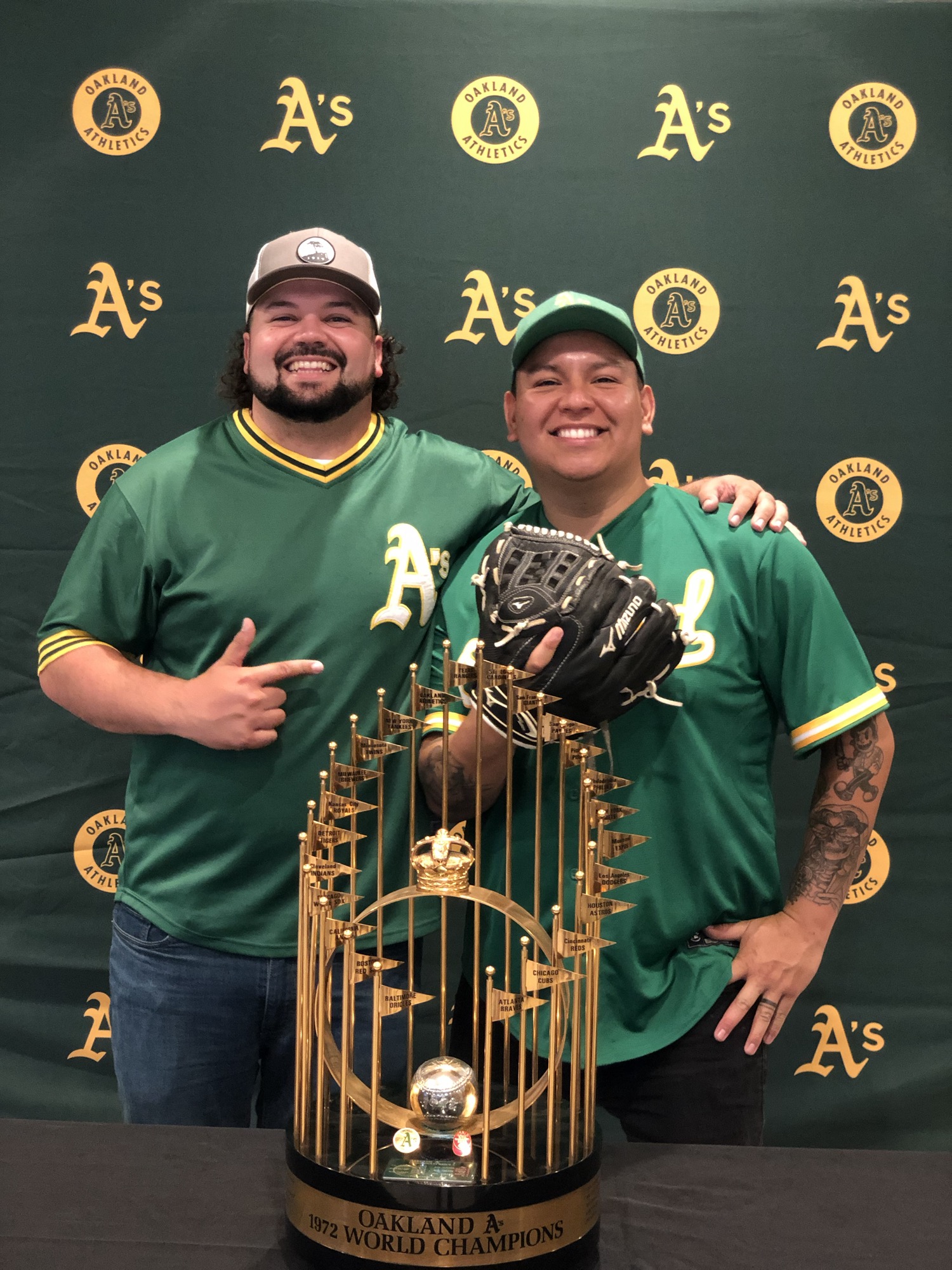 Two men wearing green and gold baseball jerseys along with green baseball hats, smile from behind the golden Oakland A's 1972 World Champions trophy.