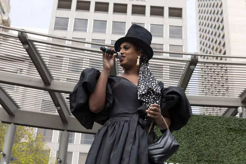 A Black trans woman with a black top hat and black dress speaks into a microphone.