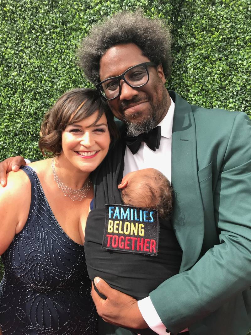 A Black man wearing glasses and a green suit with a black bow tie has his hand over the shoulder of a white woman wearing a dark blue dress while he holds a baby in a sling with a patch that reads "Families Belong Together."