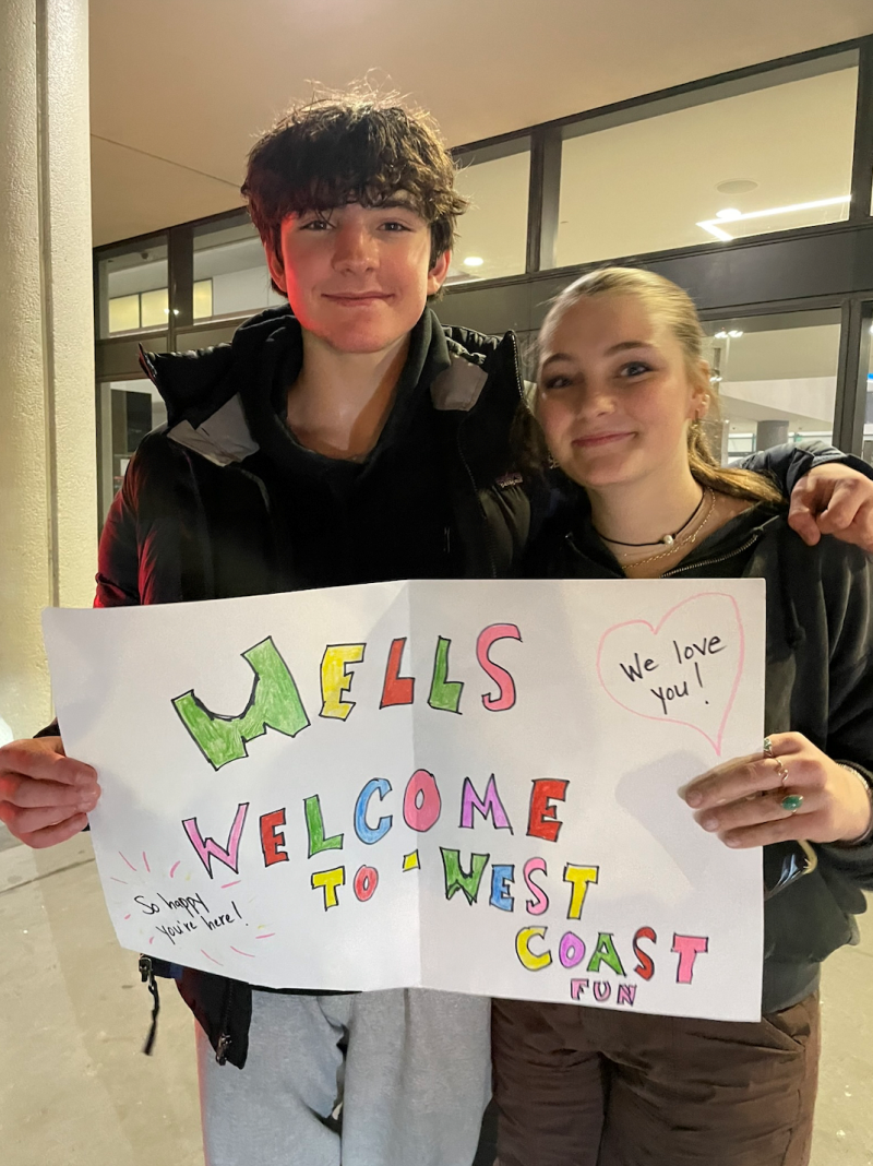 A teen boy with shaggy brown hair stands with his arm on the shoulder of a girl with blonde hair pulled back in a ponytail. They are cousins. The teens hold a sign that reads, "Wells, Welcome to West Coast fun."