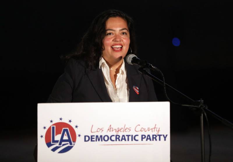A Latina woman speaks into a microphone behind a dais with a sign that reads "Los Angeles County Democratic Party."
