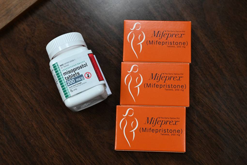 A white pill bottle with a label that reads, "misoprostol tablets 200 mcg" lies on a wooden surface. To the right, three identical orange medicine boxes read, "Mifeprez (Mifepristone) Tablets, 200 mg."