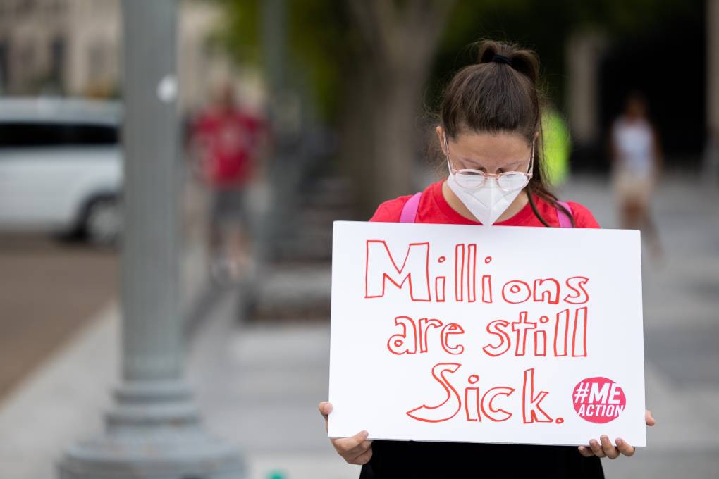 A woman with long, brown hair and eyeglasses wears a white mask and a red T-shirt. She holds a white sign with red letters that reads, "Millions are still sick. #meaction" Other protestors are seen blurred in the background. It's a protest in front of the White House.
