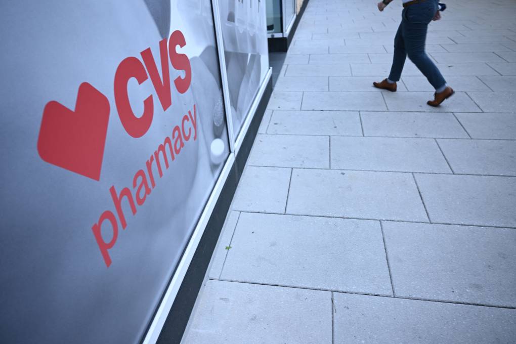 A red sign for CVS Pharmacy is displayed on the outside of its building. A person's legs are seen walking toward the entrance.