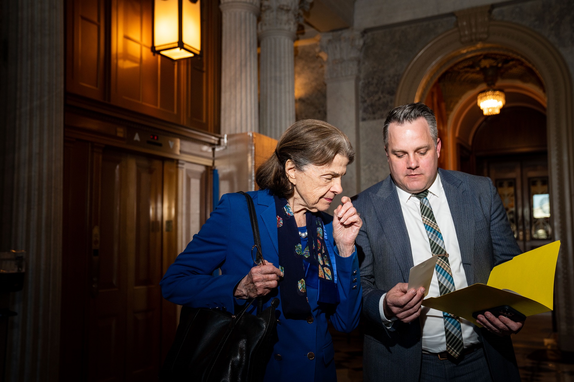 An older white woman with light, brown hair and a blue business suit stands next to another man with gray hair and a gray suit. He holds a yellow folder and is showing the woman a document inside a government building.