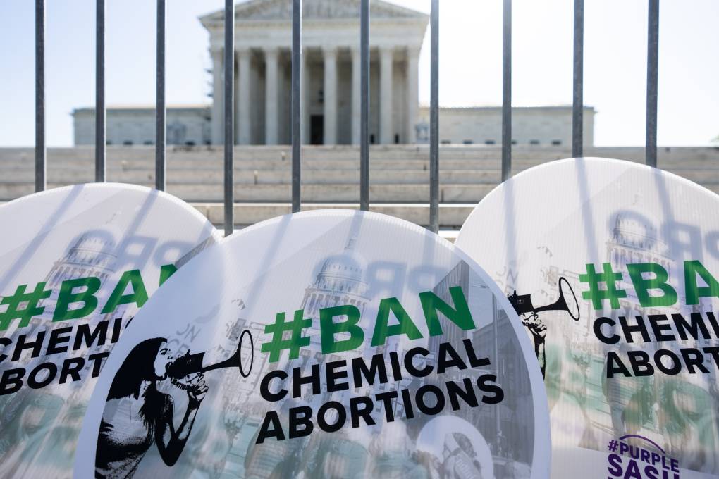 Three circular-shaped signs read "#BAN CHEMICAL ABORTIONS" and are held outside the Supreme Court, seen through bars of a gate.