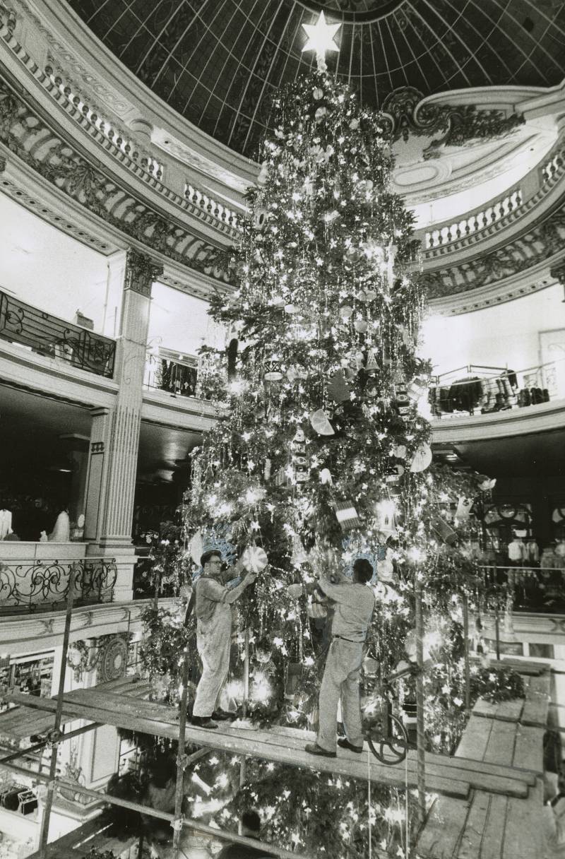 Black and white image of two people decorating a 35-foot Christmas tree. Every part of the tree is covered in glittering lights and ornaments, with an enormous star on top. 