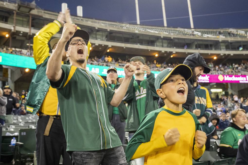 A 10-year-old boy wearing an Oakland A's baseball cap and a green and yellow baseball shirt cheers loudly alongside a huge crowd of baseball fans at a stadium.