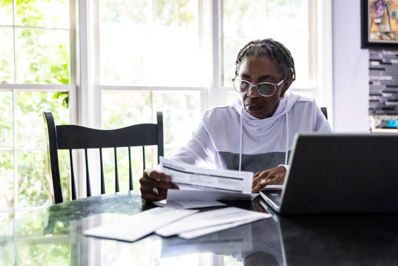 A woman sits at her kitchen table and sifts through documents, looking concerned. Next to her is her opened laptop.