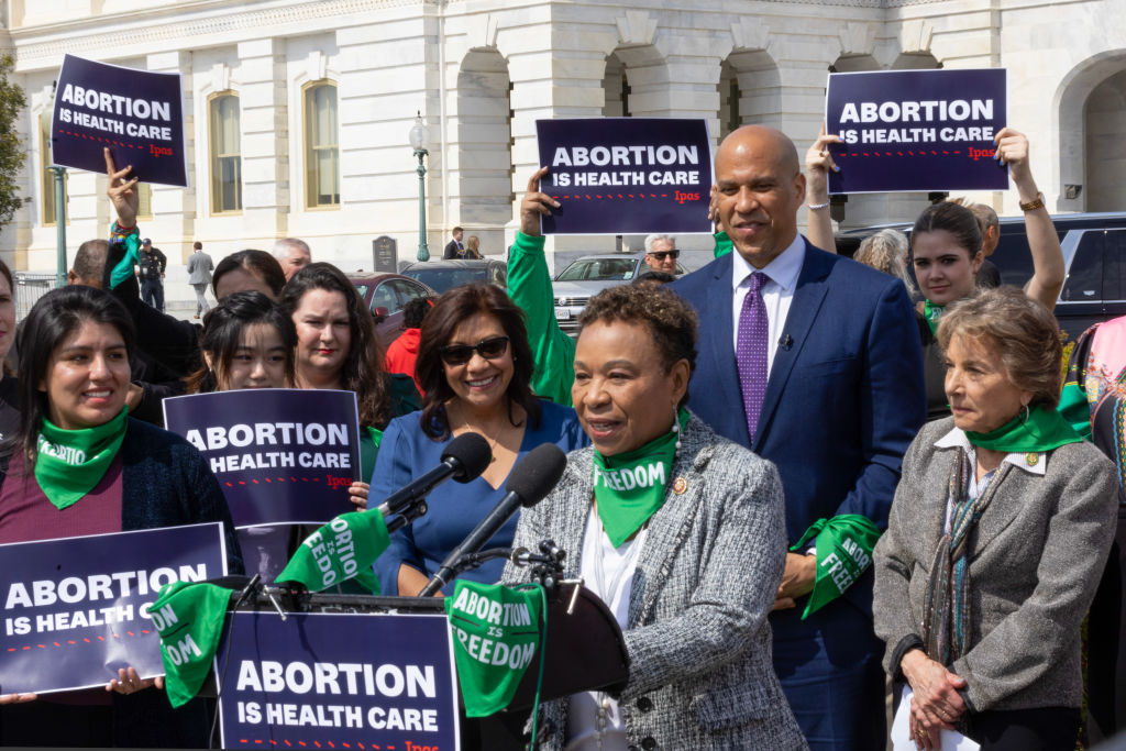 An African American woman with short hair speaks into a microphone surrounded by people holding signs that say "Abortion Is Health Care"