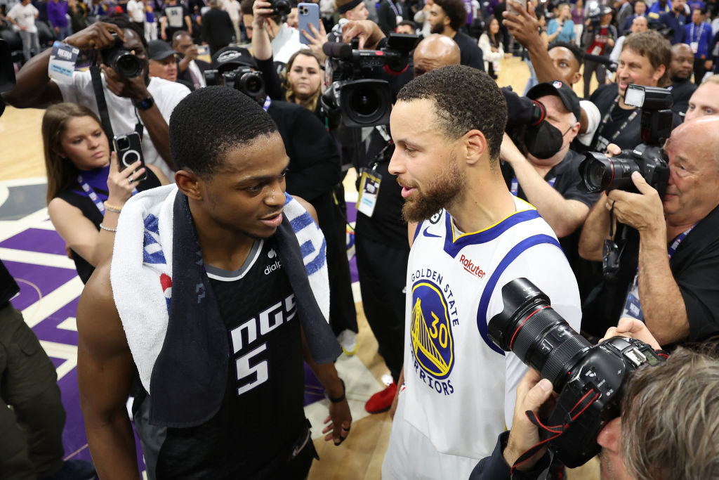 Two basketball players from opposing teams talk in front of press and fans.