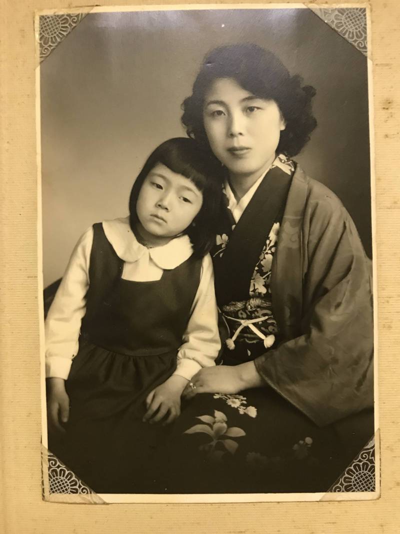 A vintage black and white photo of an Asian woman wearing a kimono and her little girl, wearing a white blouse with a butterfly collar and a dark pinafore, sitting on her lap. The woman is smiling, close-lipped, and the little girl is looking down and to the left, away from the camera.