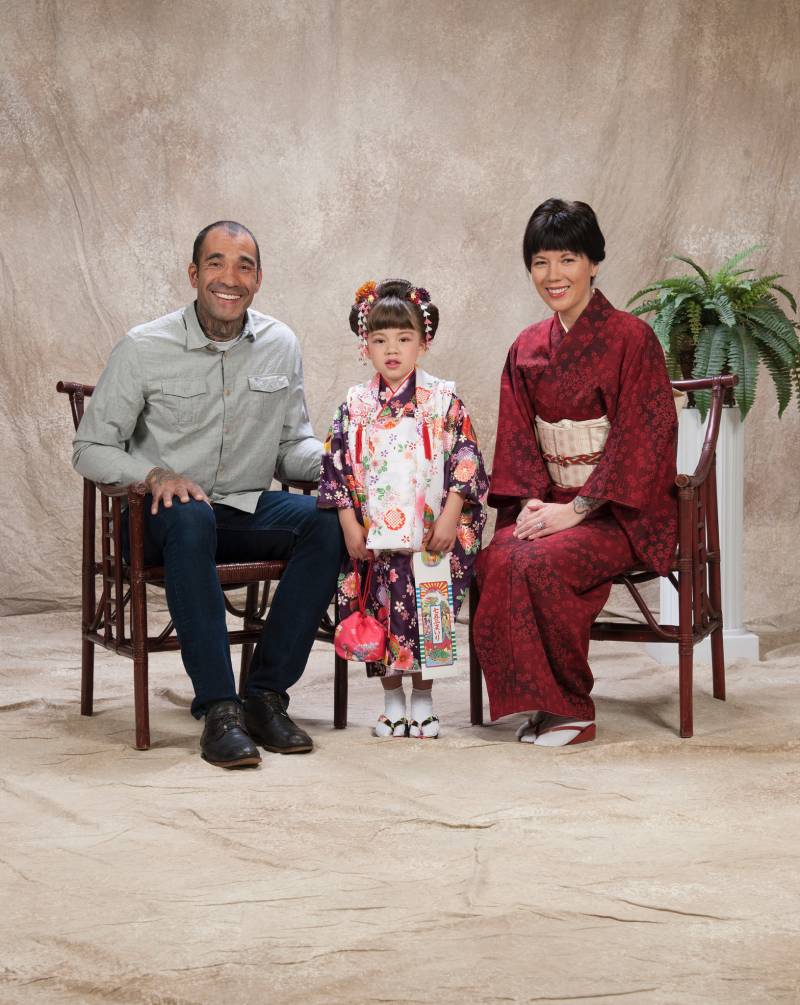 A man wearing a dress shirt, dark pants and shoes sits to the left of a small child in a dress standing in the middle and woman in a red kimono seated to the right.
