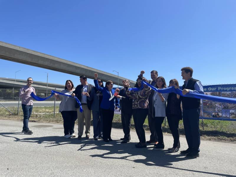 Ten people stand in a row with a freeway behind them as they cut a blue ribbon and smile.