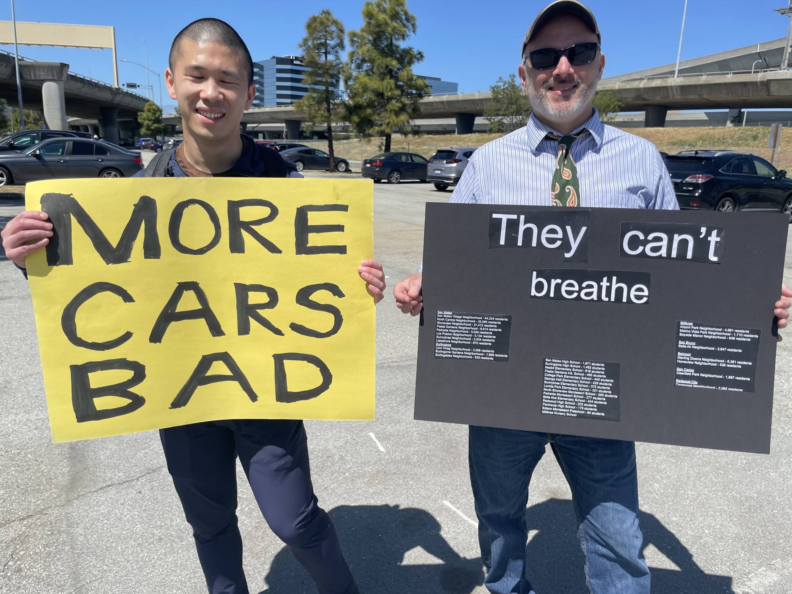 Two men, one young and Asian with a shaved head, the other middle-aged and white, with sunglasses, a baseball cap and a white beard, both smiling, hold signs that read "More Cars Bad" and "They Can't Breathe"