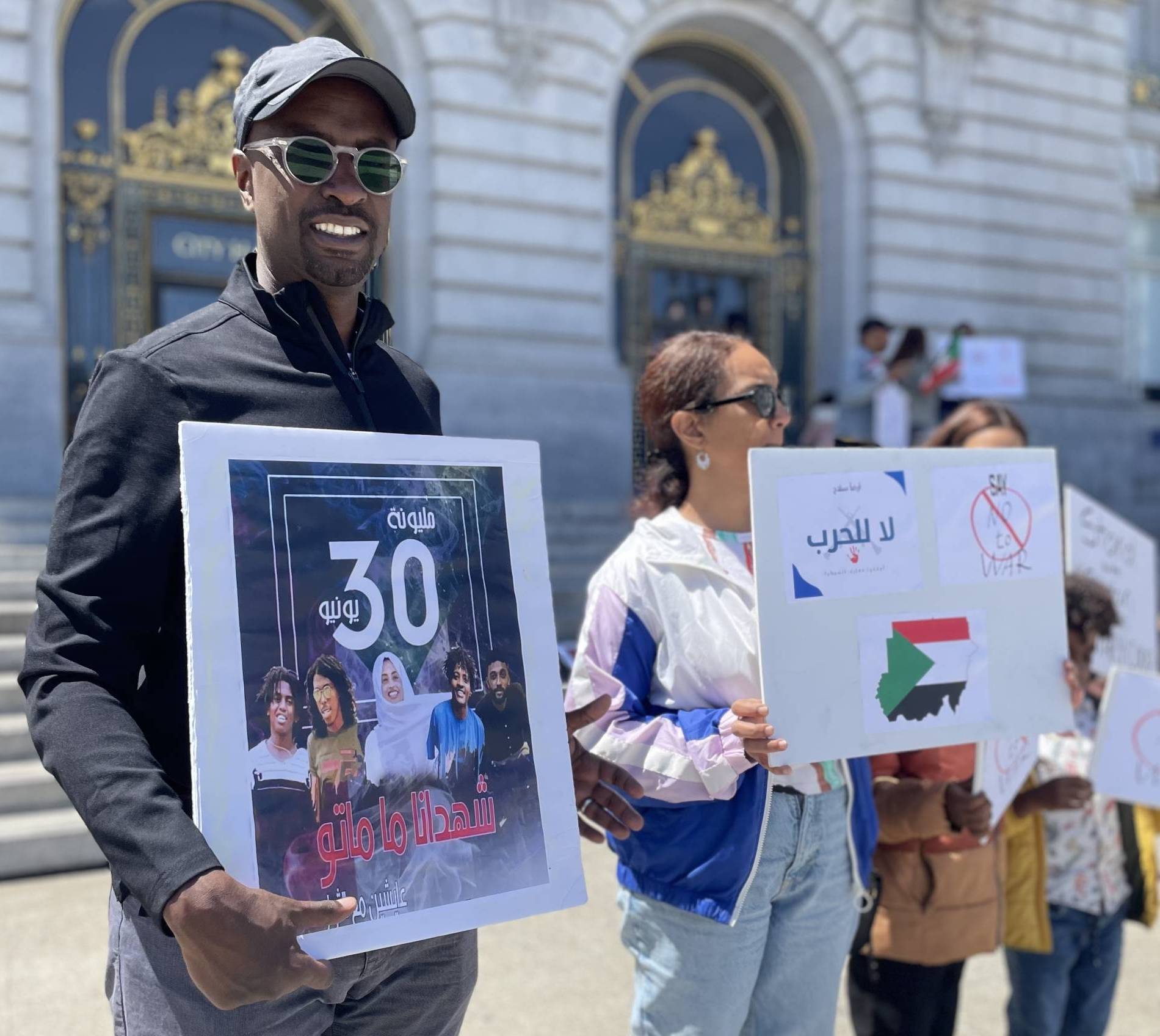 Sudanese Americans stand outside SF City Hall holding signs. 