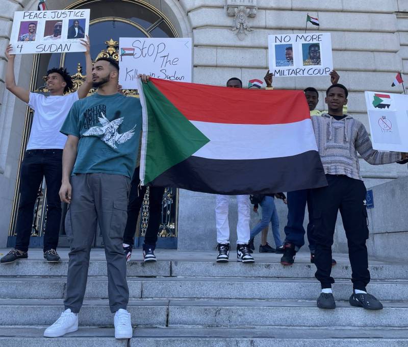 Various Sudanese American people hold signs and a large Sudanese flag on the steps of City Hall in San Francisco.