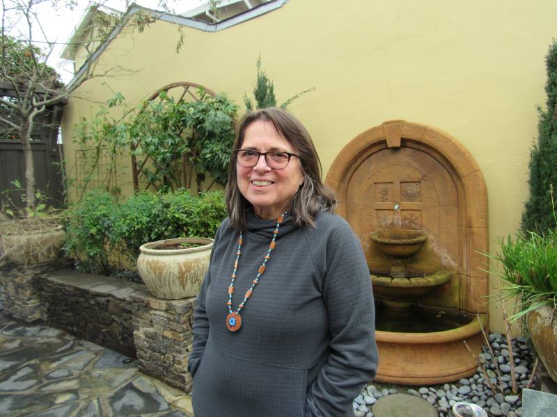 A woman wearing glasses, a necklace and gray sweater stands behind a fountain and outdoor garden.