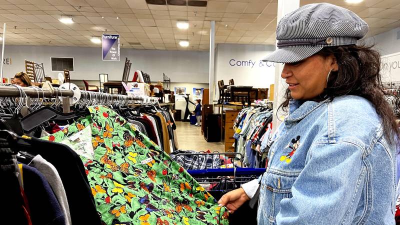 A woman with a hat and a jean jacket looks at a green patterned dress at a thrift store. 