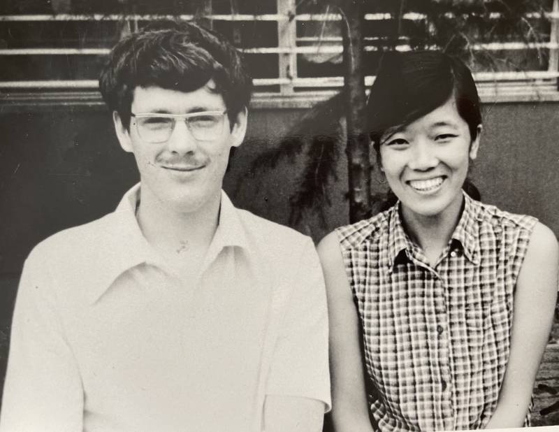 A vintage black and white photo of a white man and an Asian woman sitting next to each other, both smiling happily and wearing short-sleeved shirts.