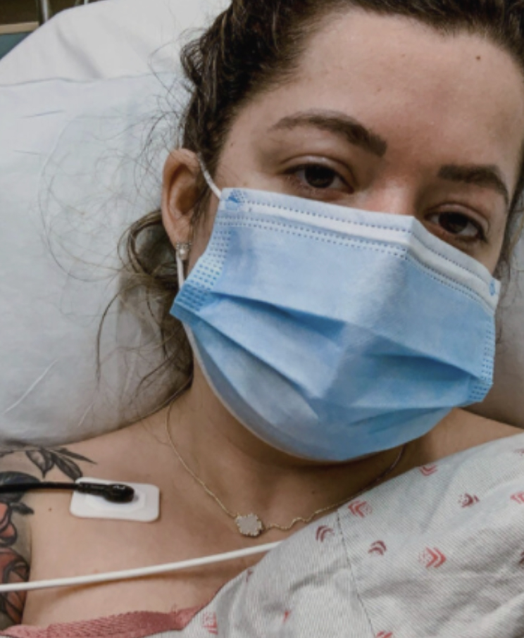 A white woman in a blue mask lies in a hospital bed with monitors stuck to her chest alongside a floral shoulder tattoo and a small gold chain with a white gemstone pendant.
