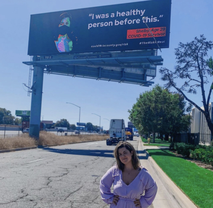 A woman stands in front of a billboard pictured in the distance. It reads, 