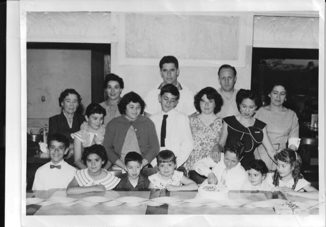 A vintage black-and-white photo of men, women and children sitting and standing in front of a table with a male child putting his hand in a cake.