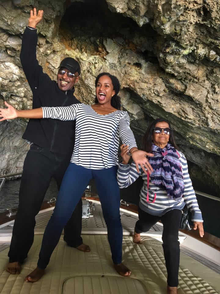 A family with an older mother and father stand on a boat with their grown-up daughter as they pose barefoot making silly faces. The boat looks as though it's inside a cave-like environment on the water.