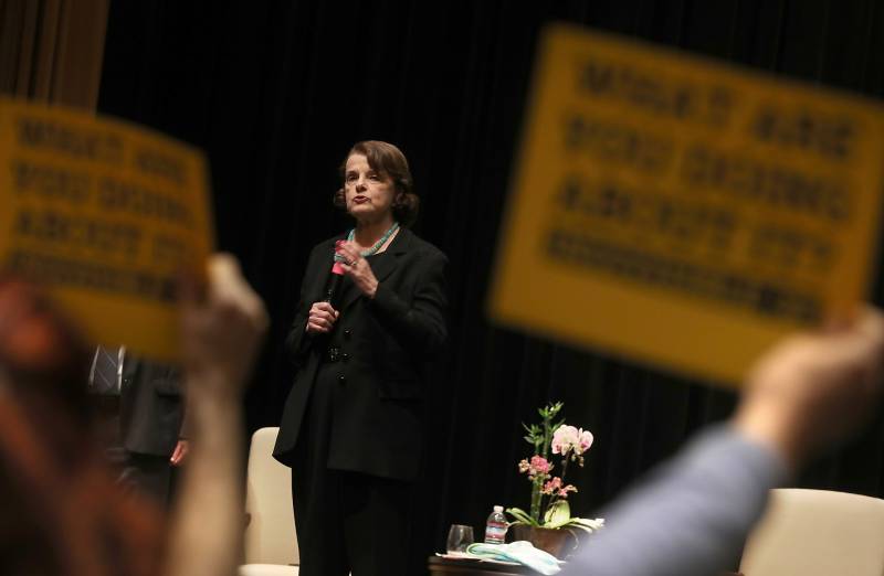 Sen. Dianne Feinstein wears a black pant suit as she speaks from a stage. In the foreground, many people's arms are seen holding black and yellow signs that are blurred.