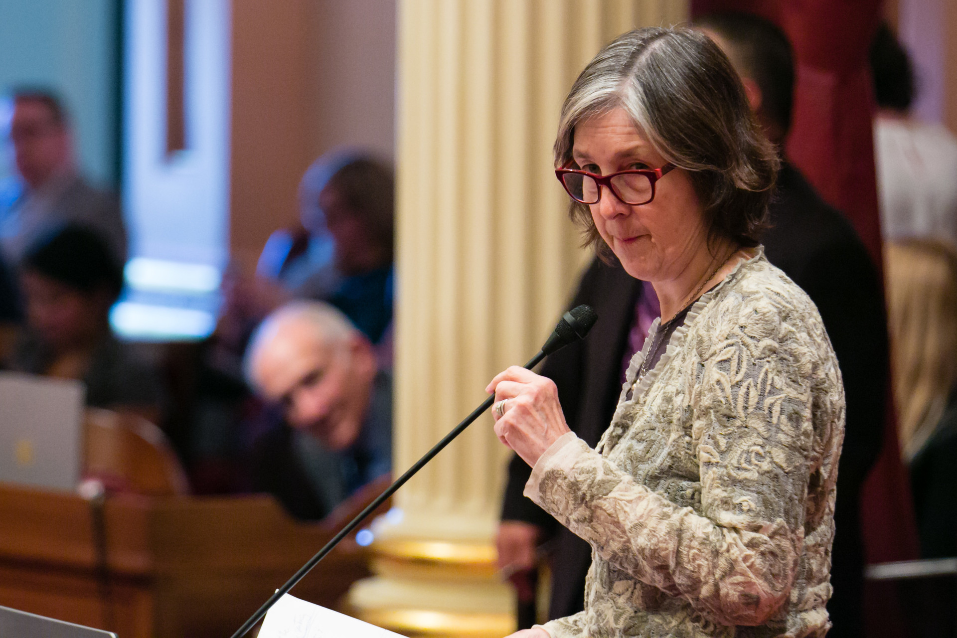 A white woman in business attire, eyeglasses and short, graying brown hair talks from a lectern inside a California government building.