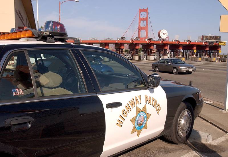 A black and white California High Patrol vehicle is parked near the Golden Gate Bridge in San Francisco. A gray car passes through the toll bridge.