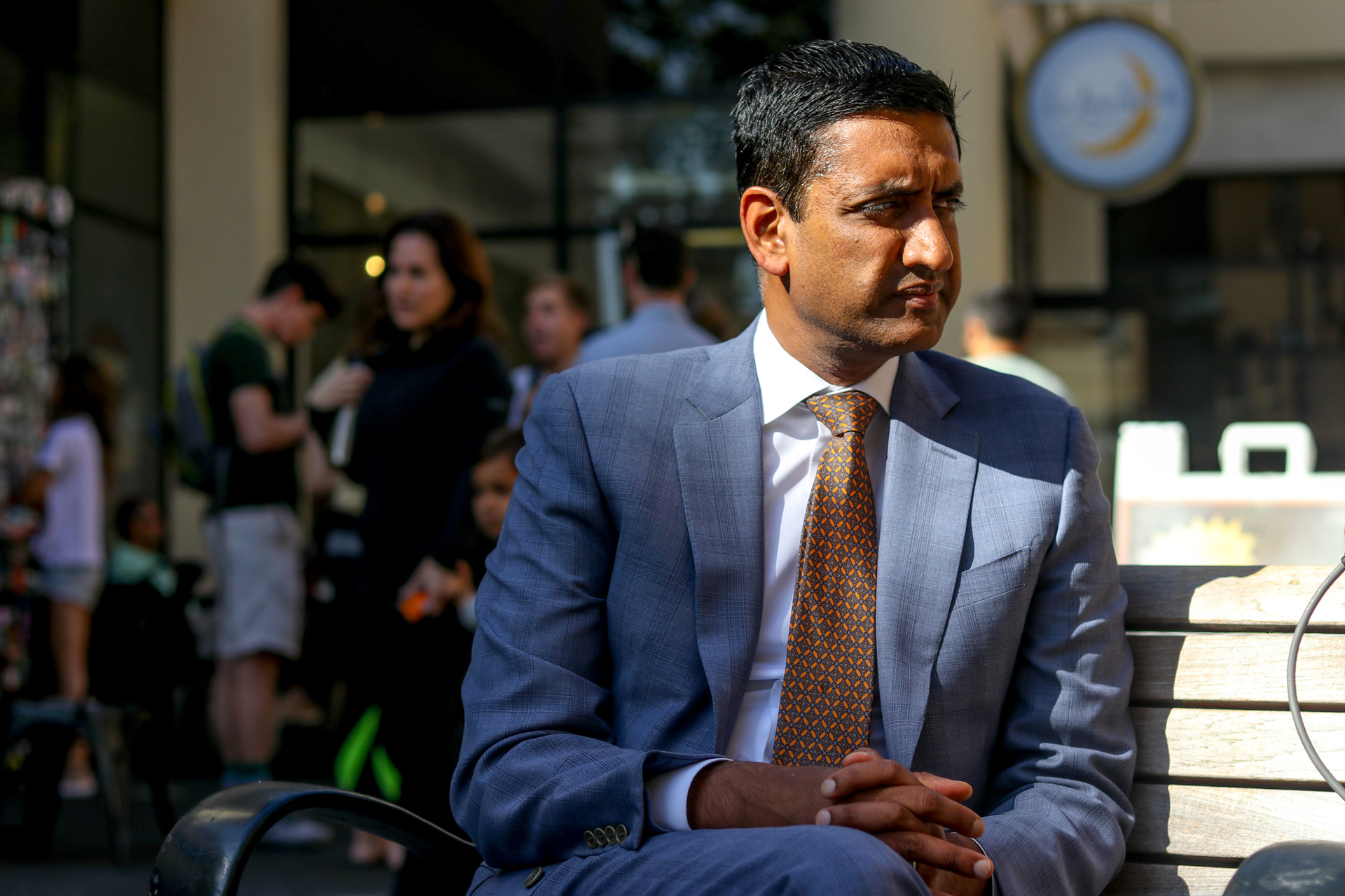 An Indian man with dark hair and eyes wears a light blue business suit and busy orange and green tie sits on a wooden bench outside. He sits crossed-legged with his arms folded on his knee. He looks to the right of the camera. Crowds of people and children are pictured behind him.