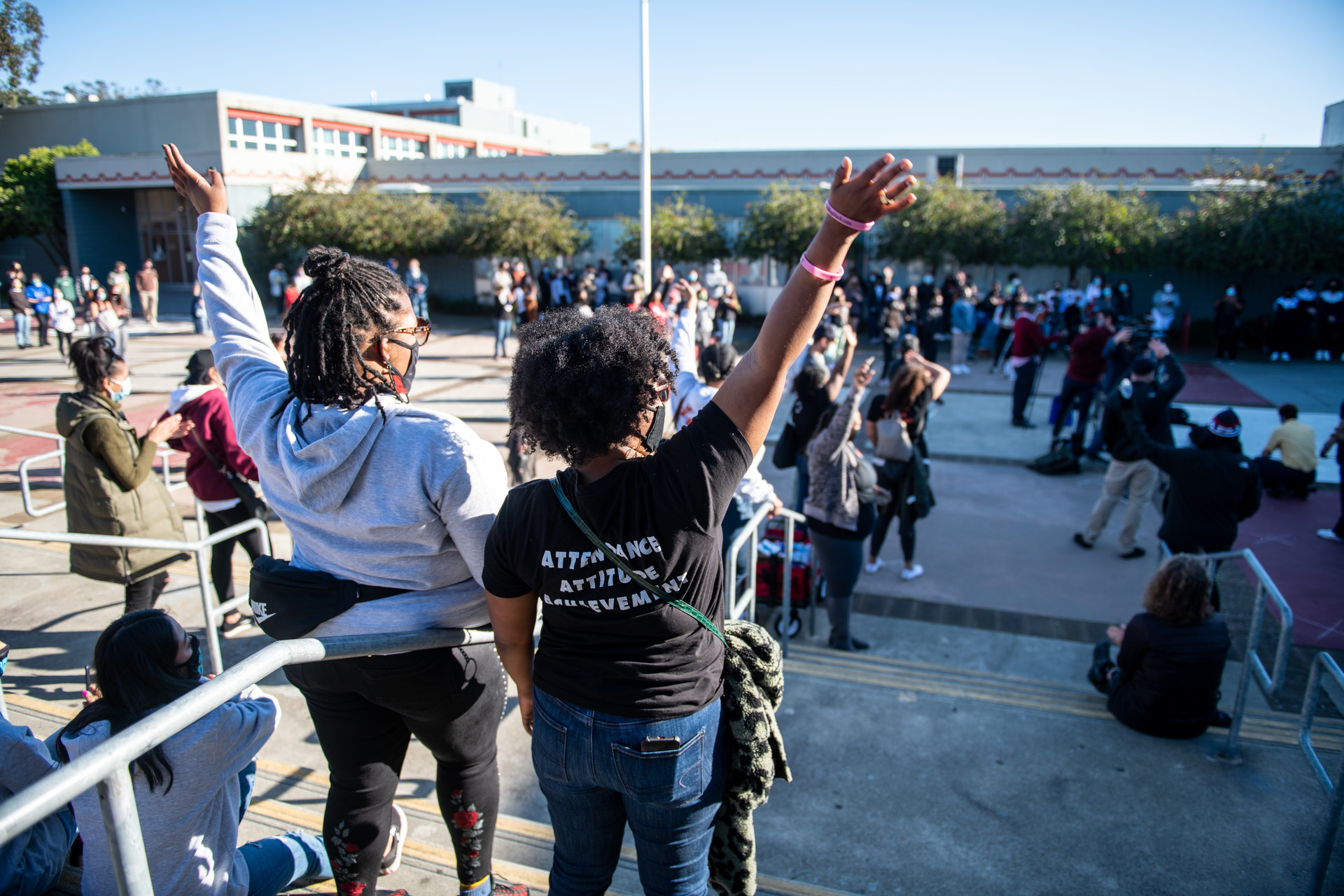Two students stand outdoors on a cement staircase with their backs to the camera and their hands raised in the air amid a crowd of other students and educators protesting at a high school campus.