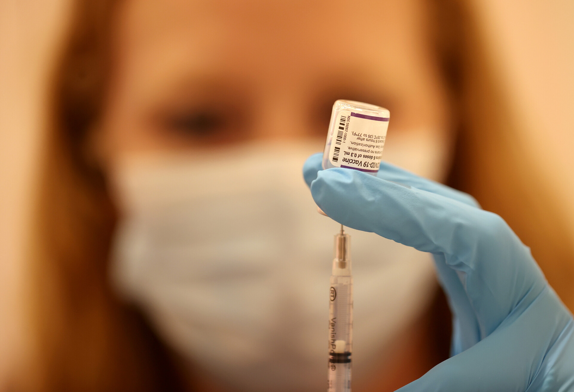 A woman with long blond hair, wearing a white face mask, fills a syringe from a small, clear vile that contains a Pfizer booster.