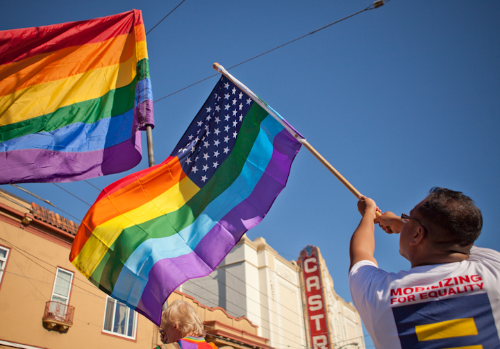 A man with a white t-shirt is seen from behind waving a Pride flag next to another Pride flag being waved with the Castro Theatre in the background.