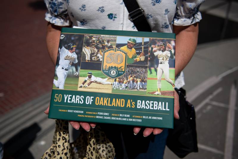 An Oakland A's baseball fan holds a special green and yellow book that chronicles 50 years of the team. She is wearing a white and blue floral top.