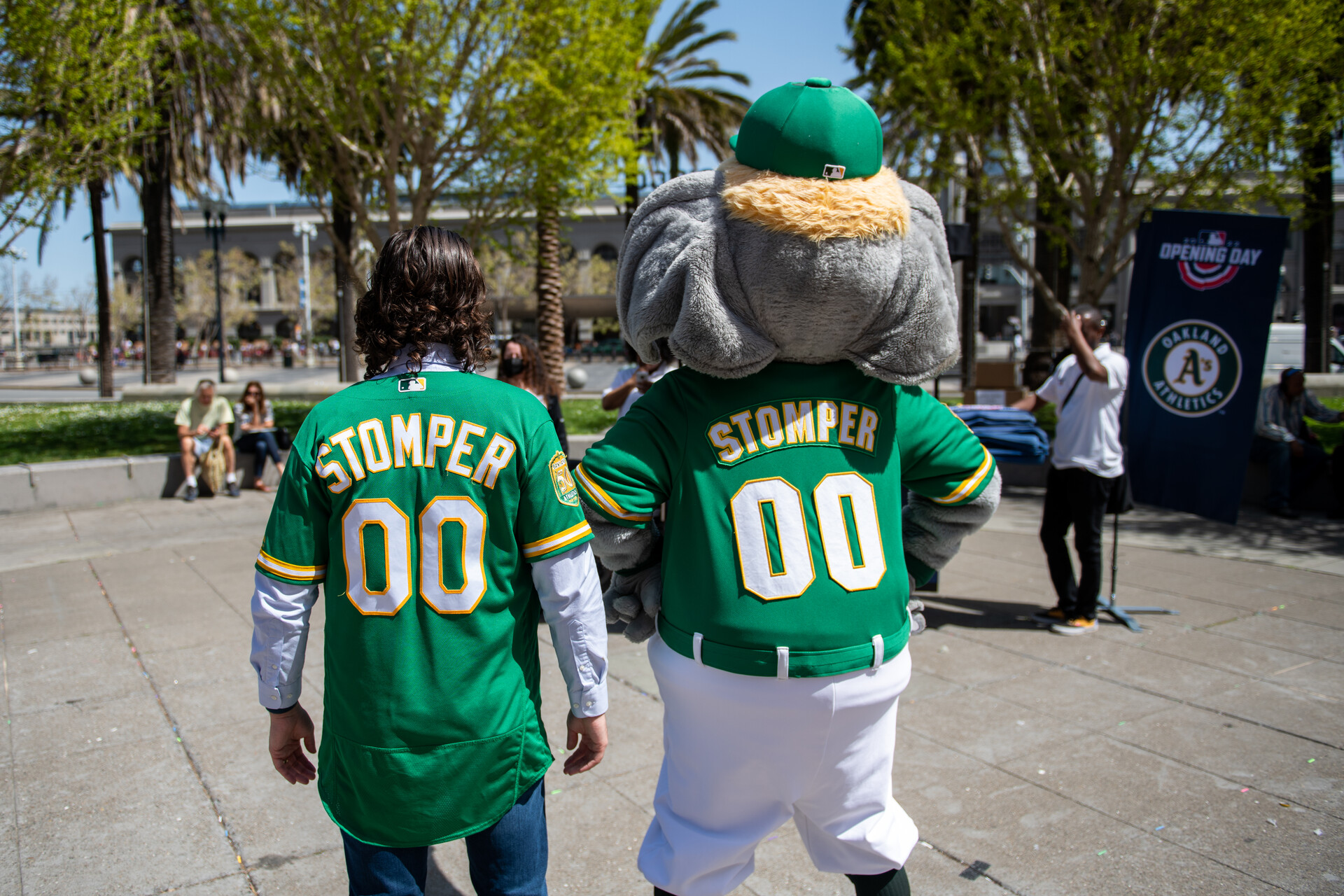 An Oakland Athletics baseball fan wears the team's green and gold jersey. The fan poses for a photo with their back toward the camera and is standing next to the team's mascot, Stomper, who is wearing a baseball uniform. Stomper is an elephant.