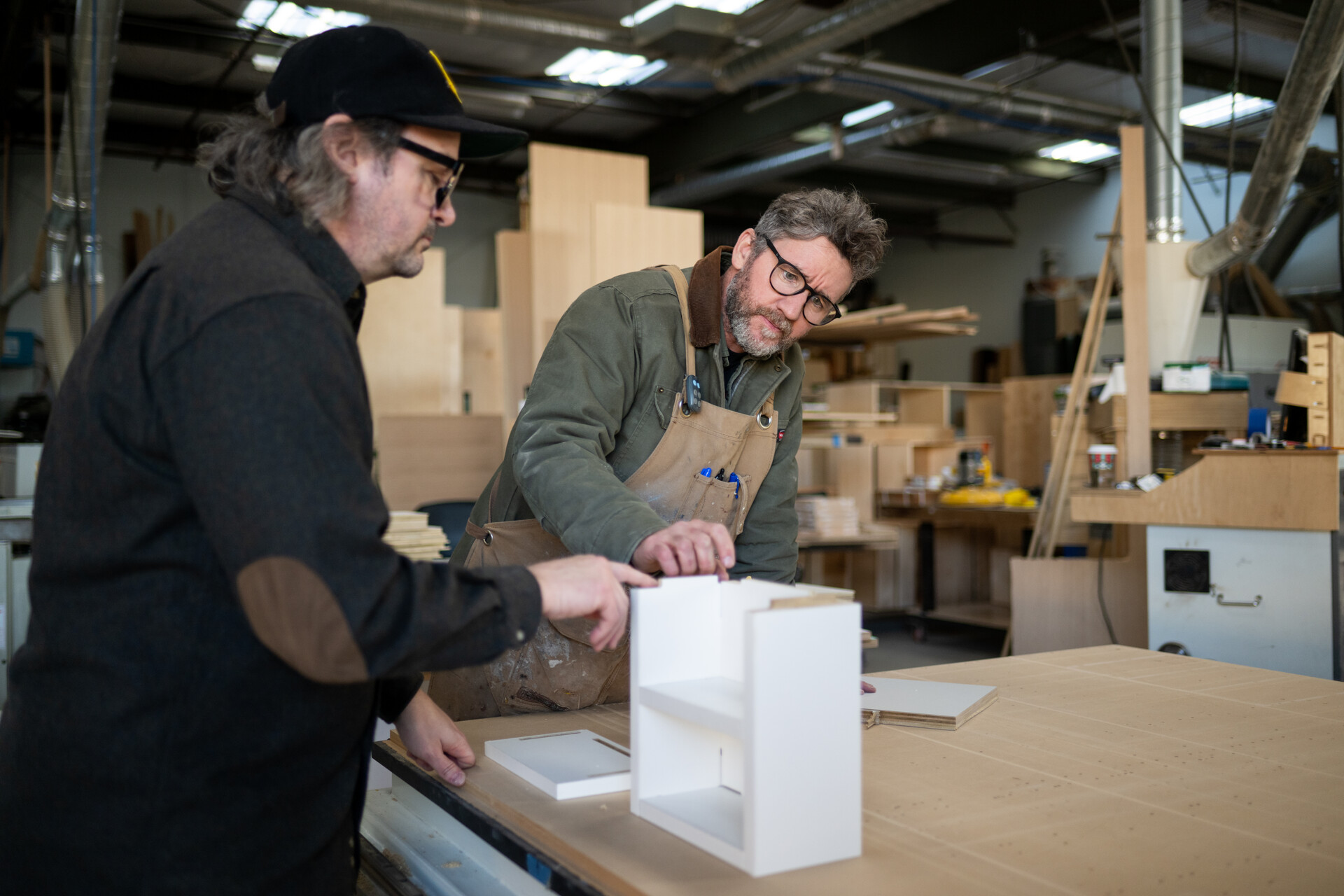 Two white men with glasses talk over a white box in process of completion in a furniture store with carpentry equipment around them.