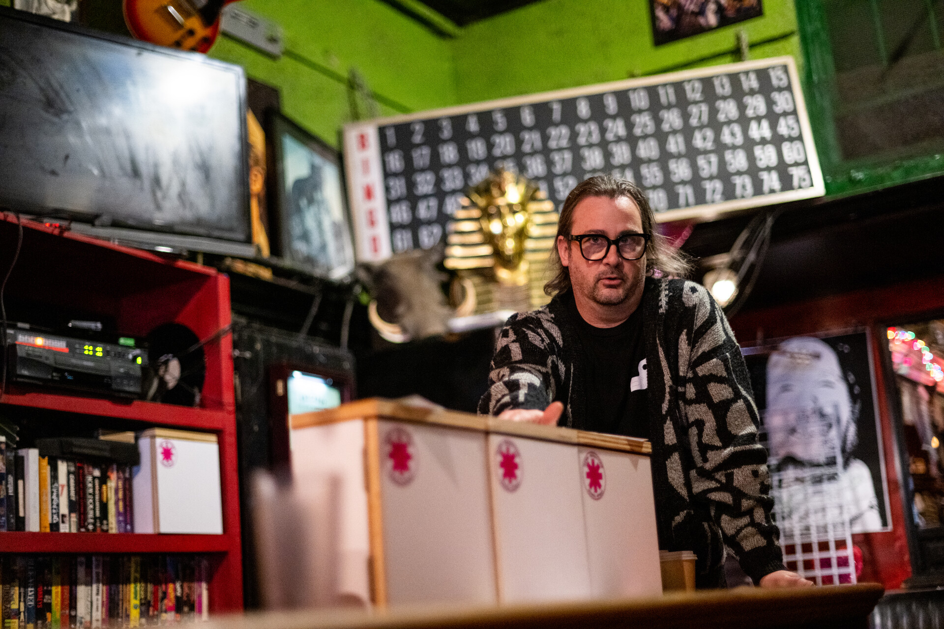 A white man with glasses looks at the camera as he gestures toward three white boxes with asterisk logos on them lined up next to each other in a bar.