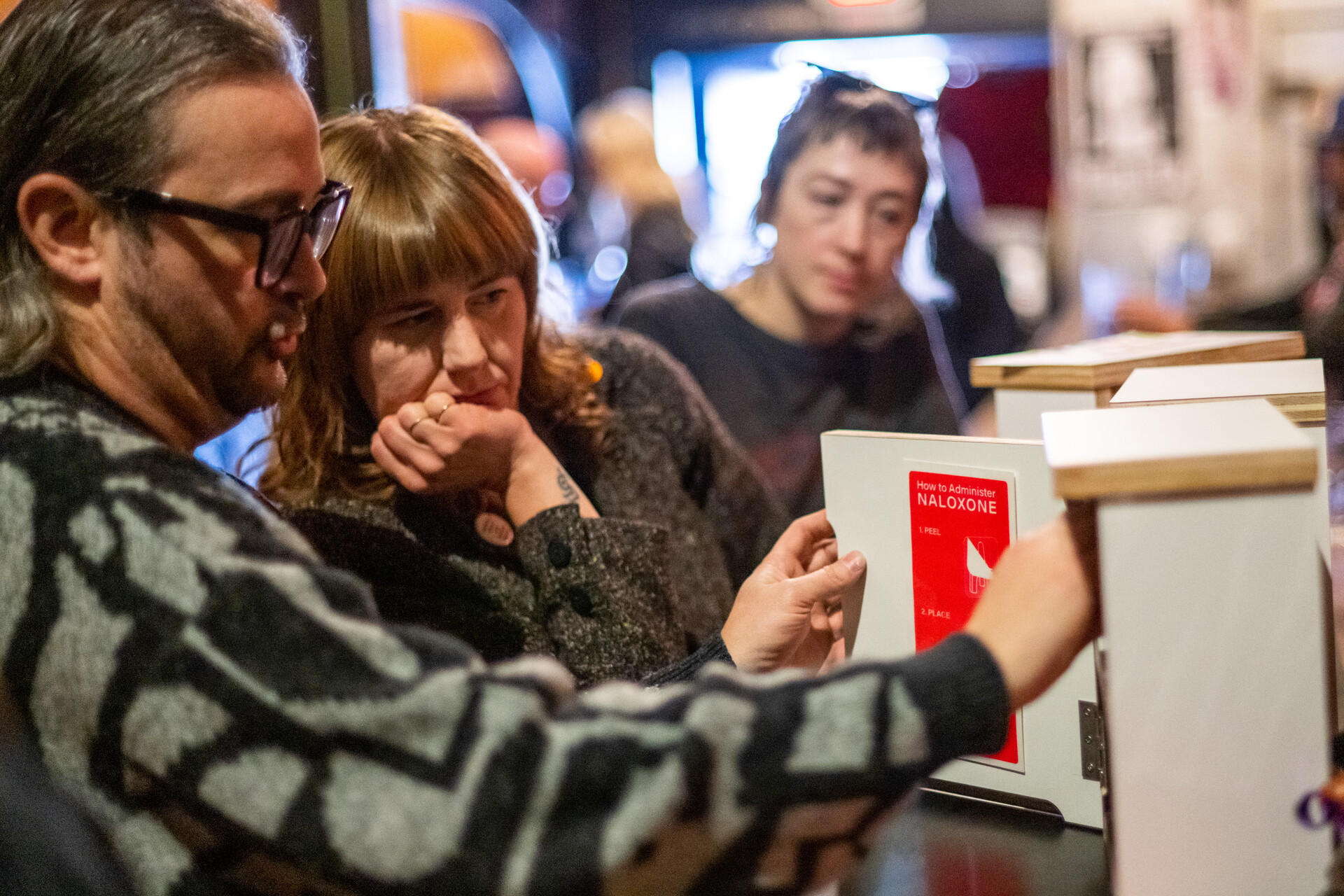 A white man with glasses and a sweater opens a white wooden box with "How to Administer Naloxone" written in red on the inside of the box lid, as two women look on from the man's left.
