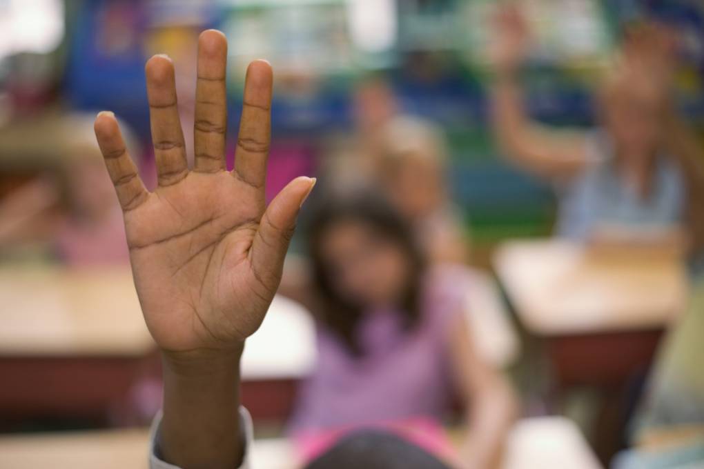 A small child's hand is raised in a classroom setting and is seen in focus. Blurred students are pictured behind the raised hand.