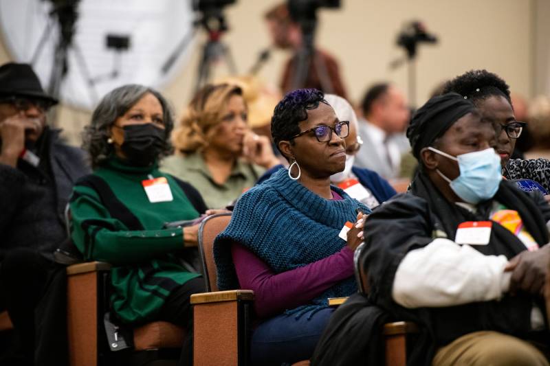 Rows of men and women sit in attendance listening intently to speakers during a California Reparations Task Force meeting. Many video cameras on tripods can be seen in the background.