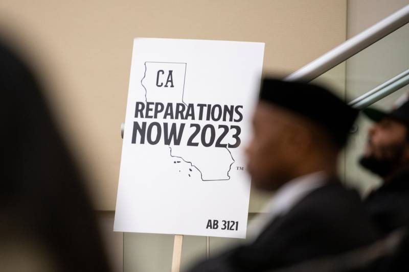 A white sign with black lettering reads "Reparations Now 2023" at a California Reparations Task Force meeting in Sacramento, the state's capital.
