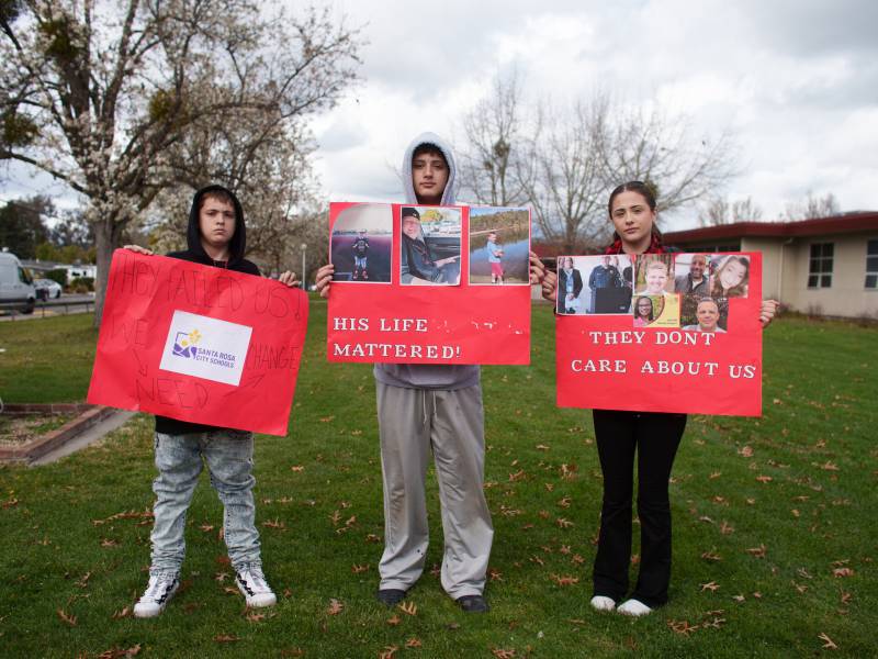 Three young people appearing to be high school aged stand equal distance apart, posing for the camera with protest signs they made, on red paper with the visible words 'his life mattered' and 'they don't care about us'